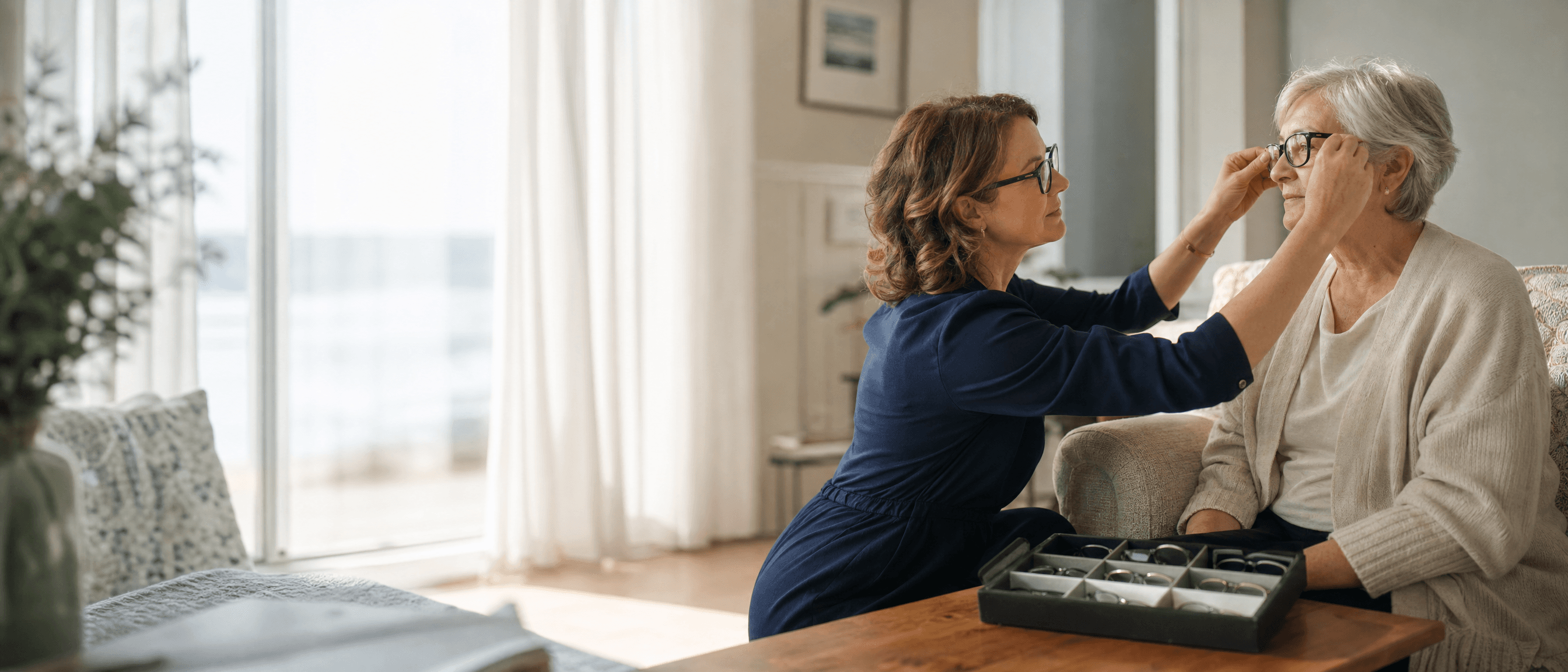 An optician fitting a pair of glasses for a seated patient in her living room